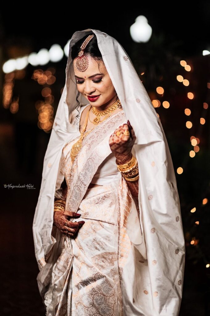 A bride wearing a white saree,smiling and looking down.